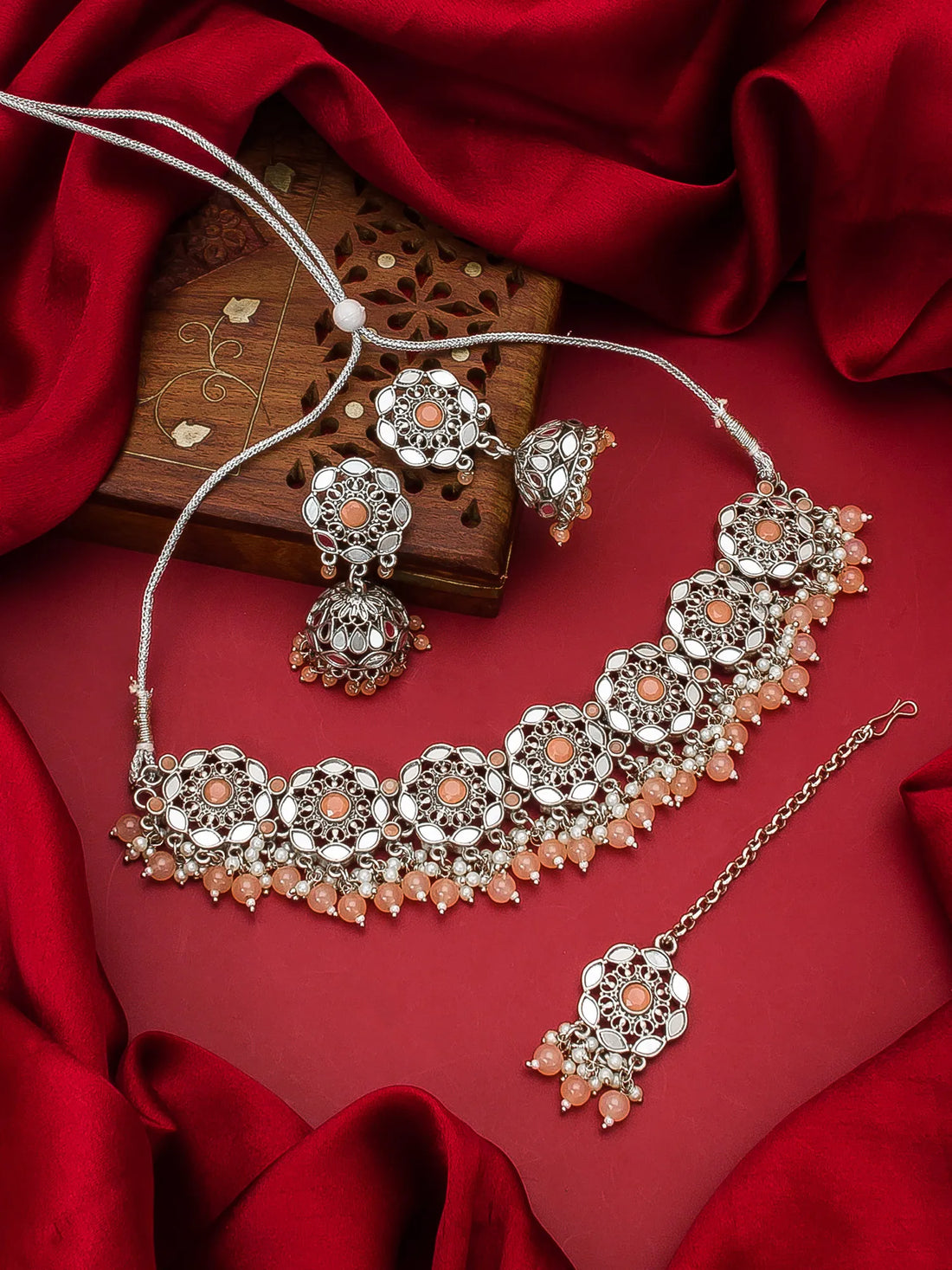 Silver-toned Indian jewelry set featuring a choker necklace, matching earrings, and a maang tikka with peach-colored stones, displayed on a red satin fabric background.