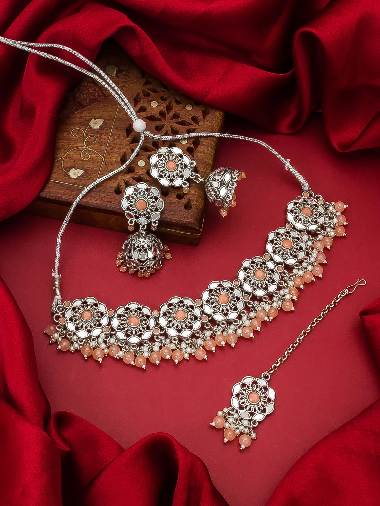 Silver-toned Indian jewelry set featuring a choker necklace, matching earrings, and a maang tikka with peach-colored stones, displayed on a red satin fabric background.