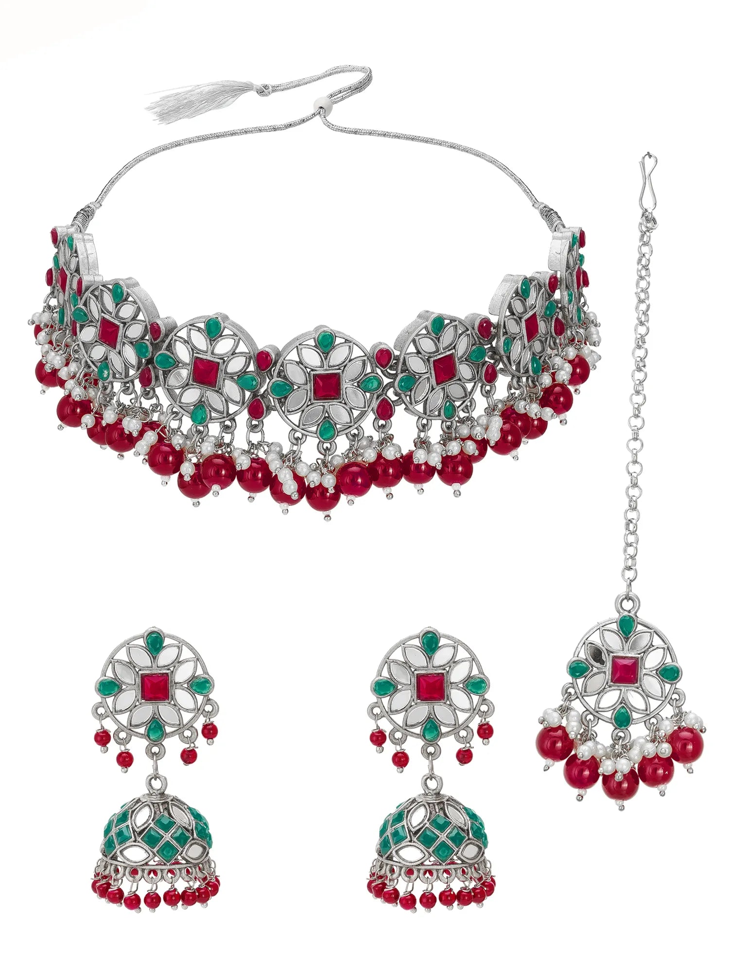 A traditional Indian jewelry set featuring a silver-toned choker necklace, matching jhumka earrings, and a maang tikka, embellished with red stones, green stones, and red pearl beads, shown on a plain white background.
