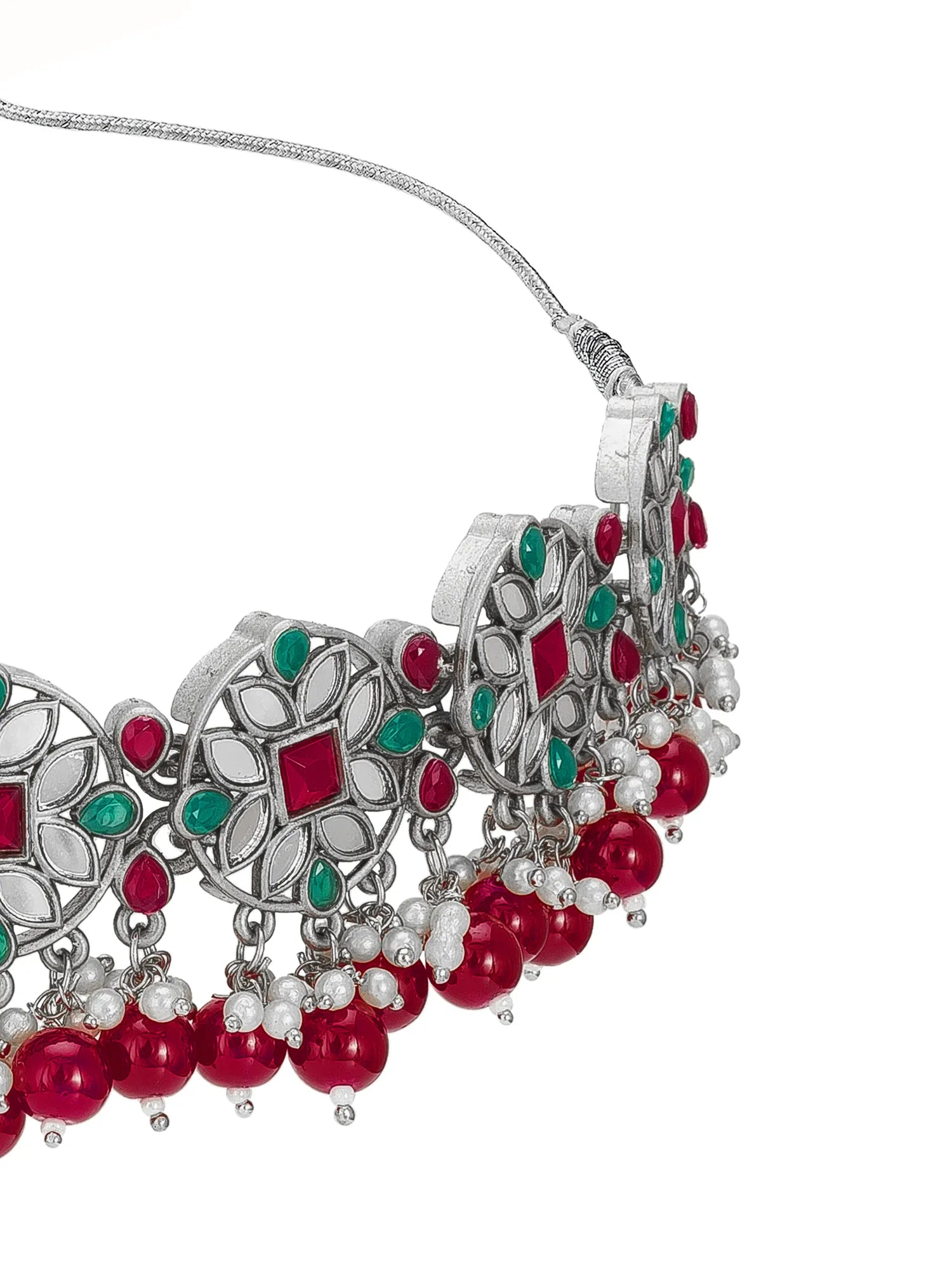 A close-up of a silver-toned statement necklace featuring intricate filigree patterns, red gemstone accents, and dangling red beads with small white pearl-like beads, shown against a plain white background.