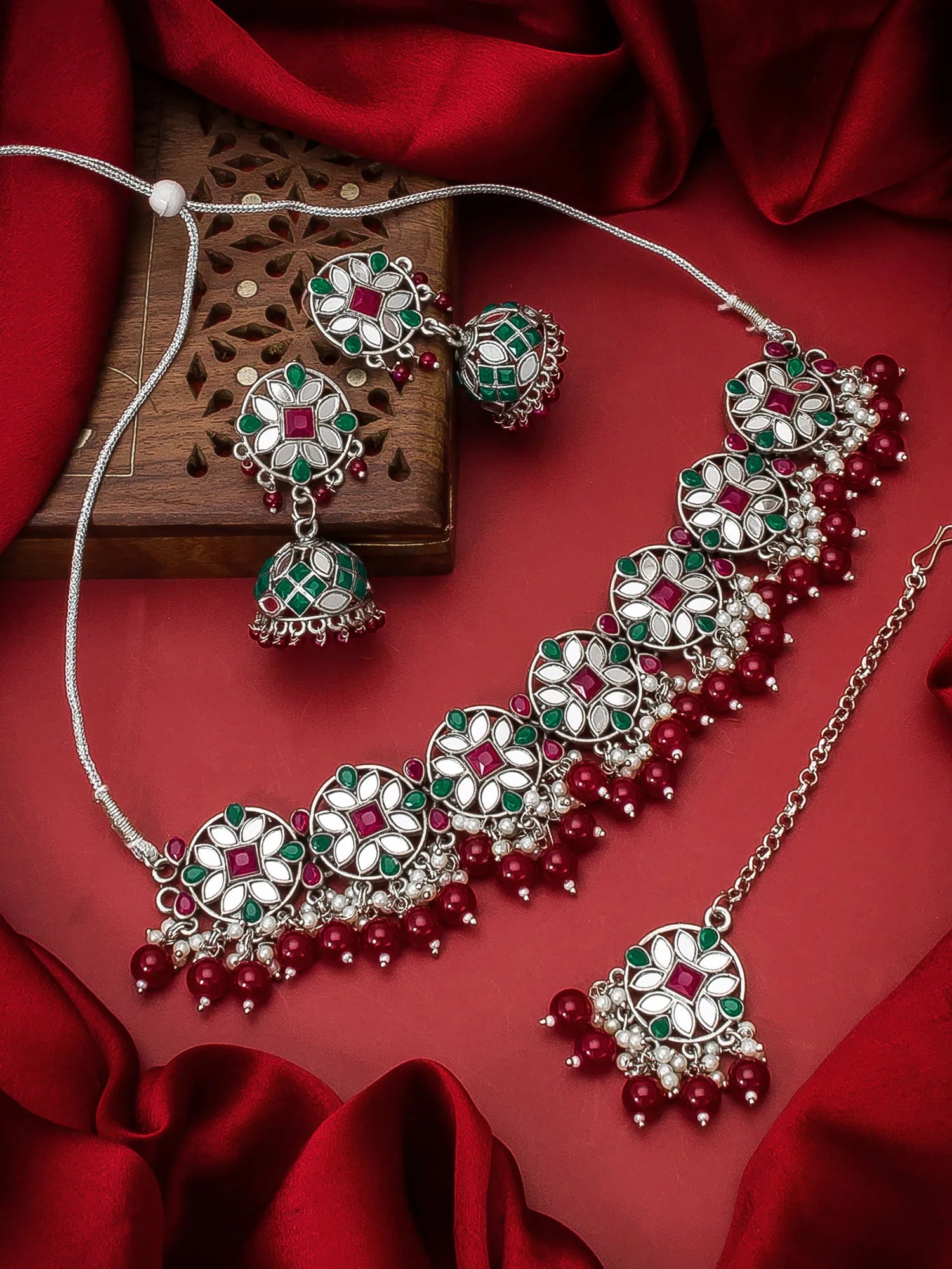 A traditional Indian jewelry set featuring a silver-toned necklace, matching earrings, and a pendant with green and white floral motifs and red bead accents, displayed on a red satin fabric background.