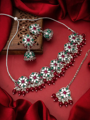 A traditional Indian jewelry set featuring a silver-toned necklace, matching earrings, and a pendant with green and white floral motifs and red bead accents, displayed on a red satin fabric background.