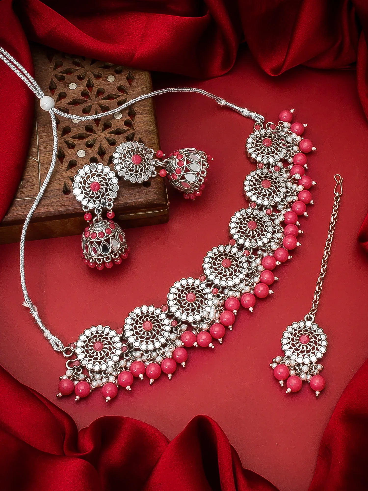 A traditional Indian jewelry set featuring a silver-toned necklace, earrings, and a matching maang tikka with pink beads and red stone centers, arranged on a red satin fabric background with a wooden box.