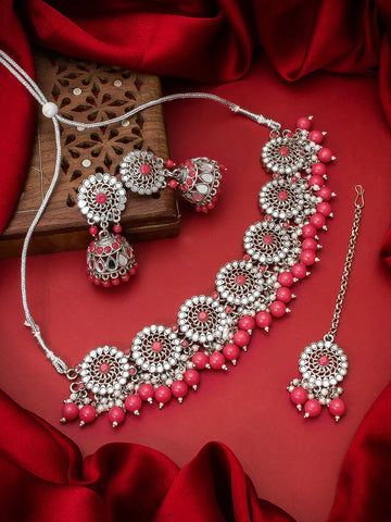 A traditional Indian jewelry set featuring a silver-toned necklace, earrings, and a matching maang tikka with pink beads and red stone centers, arranged on a red satin fabric background with a wooden box.