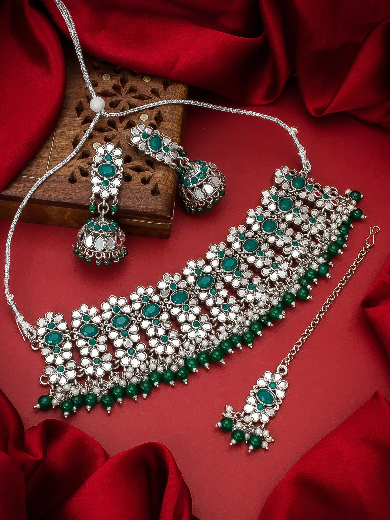 A silver-toned Indian jewelry set featuring a choker necklace, matching earrings, and a pendant, embellished with green stones and white stones, displayed on a red satin fabric background.