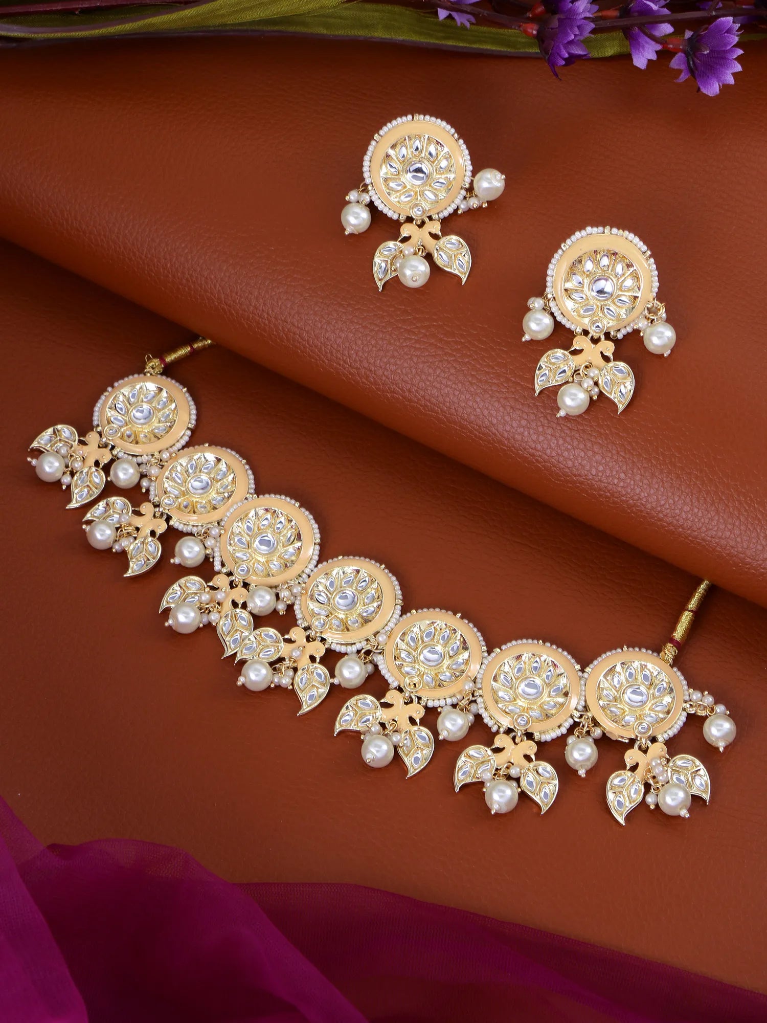 Gold-toned Indian bridal jewelry set featuring a choker necklace, matching earrings, and a maang tikka, embellished with clear crystals and white faux pearls, displayed on a brown textured leather background.