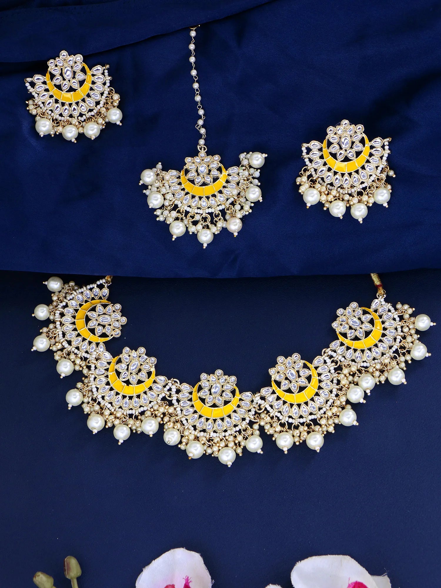 Gold-plated traditional Indian jewelry set featuring a necklace, earrings, and a maang tikka, embellished with white faux pearls and bright yellow crescent-shaped inlays, displayed on a dark navy blue fabric background.