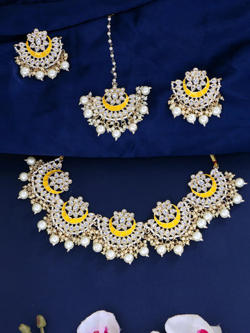 Gold-plated traditional Indian jewelry set featuring a necklace, earrings, and a maang tikka, embellished with white faux pearls and bright yellow crescent-shaped inlays, displayed on a dark navy blue fabric background.