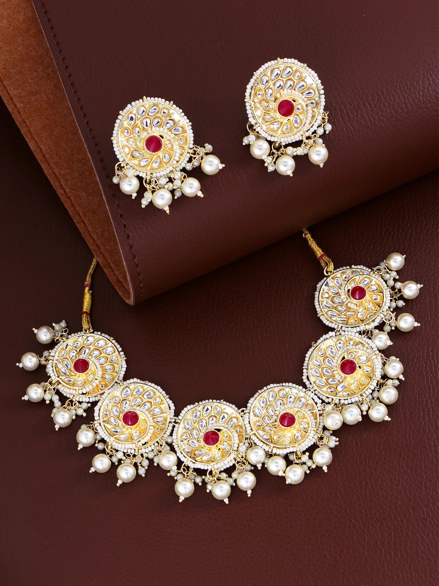 Gold-plated Indian bridal jewelry set featuring a necklace, earrings, and a maang tikka, decorated with white faux pearls, red stones, and intricate gold filigree, displayed on a brown leather background.