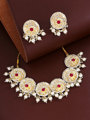 Gold-plated Indian bridal jewelry set featuring a necklace, earrings, and a maang tikka, decorated with white faux pearls, red stones, and intricate gold filigree, displayed on a brown leather background.