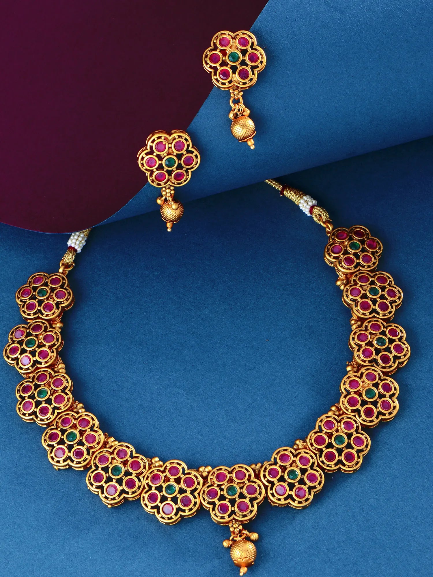 Gold-plated traditional Indian jewelry set featuring a necklace and matching earrings, embellished with pinkish-red and green stones in a floral pattern, displayed on a blue and purple background.