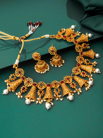 Gold-toned traditional Indian jewelry set featuring a choker necklace, matching earrings, and a beaded tassel, decorated with red stones and white pearl-like beads, displayed on a dark green background.
