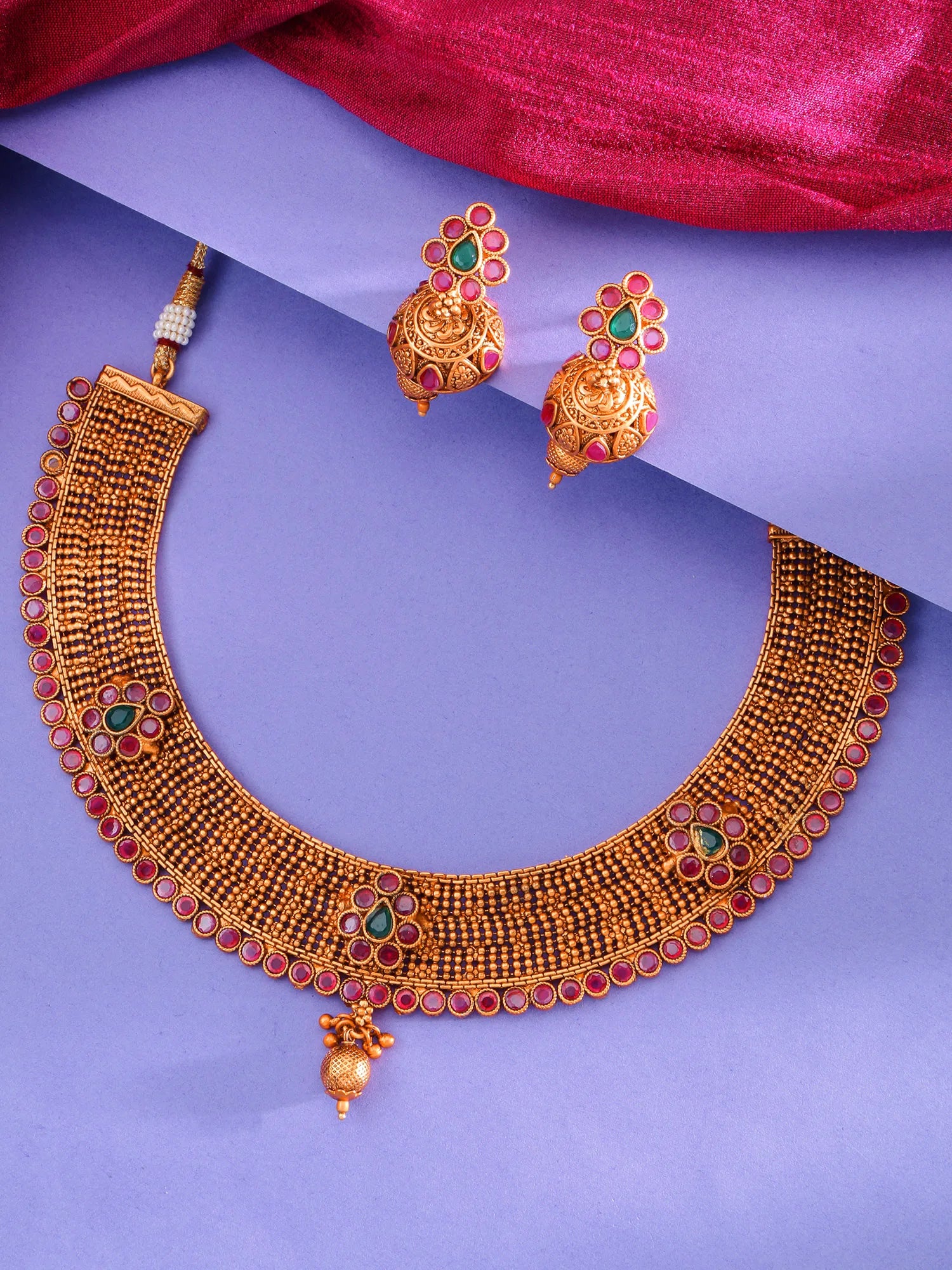 Gold-toned traditional necklace and matching earrings with red and green stone accents, arranged on a purple background with a draped magenta fabric at the top.
