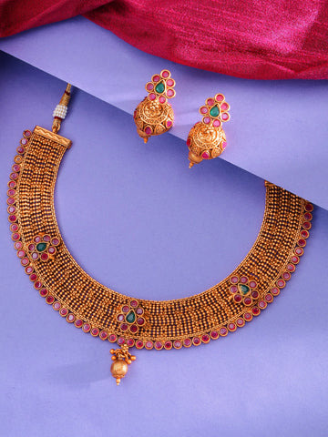 Gold-toned traditional necklace and matching earrings with red and green stone accents, arranged on a purple background with a draped magenta fabric at the top.