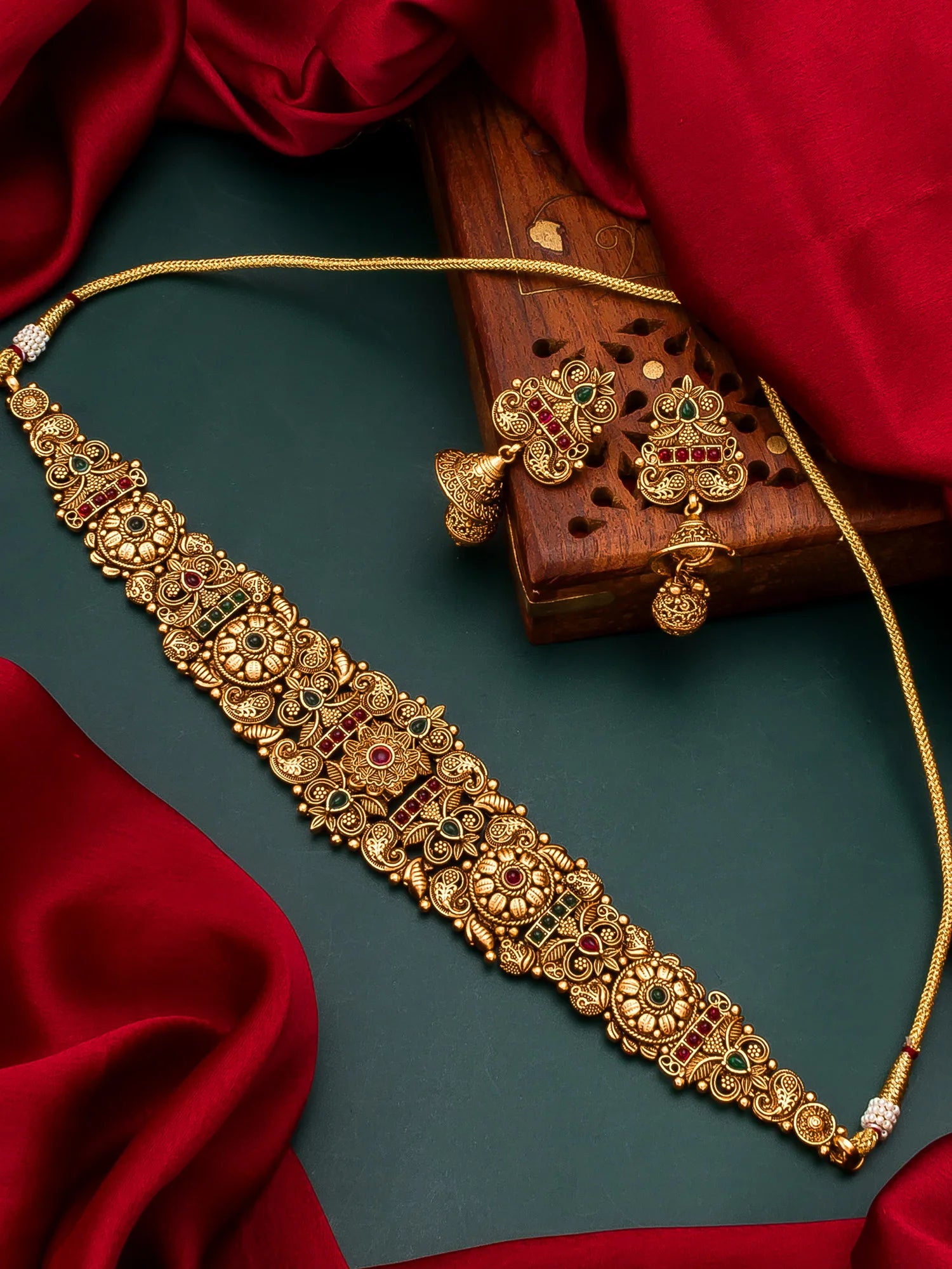An ornate gold-toned jewelry set laid out on a dark teal surface with draped red fabric and a carved wooden box. The set includes a long necklace, a matching bracelet, and dangling earrings with detailed floral motifs and small red and green stone accents.
