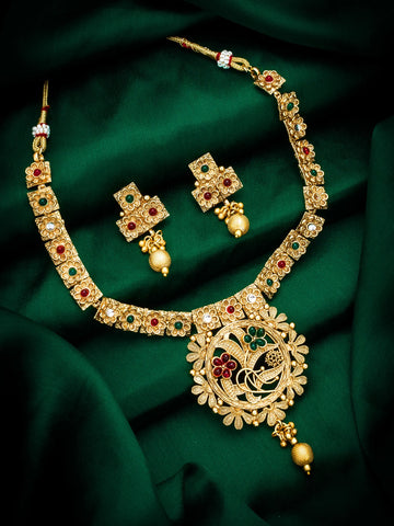 An ornate gold jewelry set featuring a necklace and matching earrings, decorated with red and green stones and gold-toned details, displayed on a draped dark green fabric background.