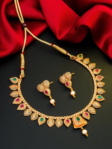Gold-toned traditional necklace and matching drop earrings with leaf-shaped motifs, red and green stone accents, and pearl-like dangling beads, arranged on a dark surface with red fabric in the background.