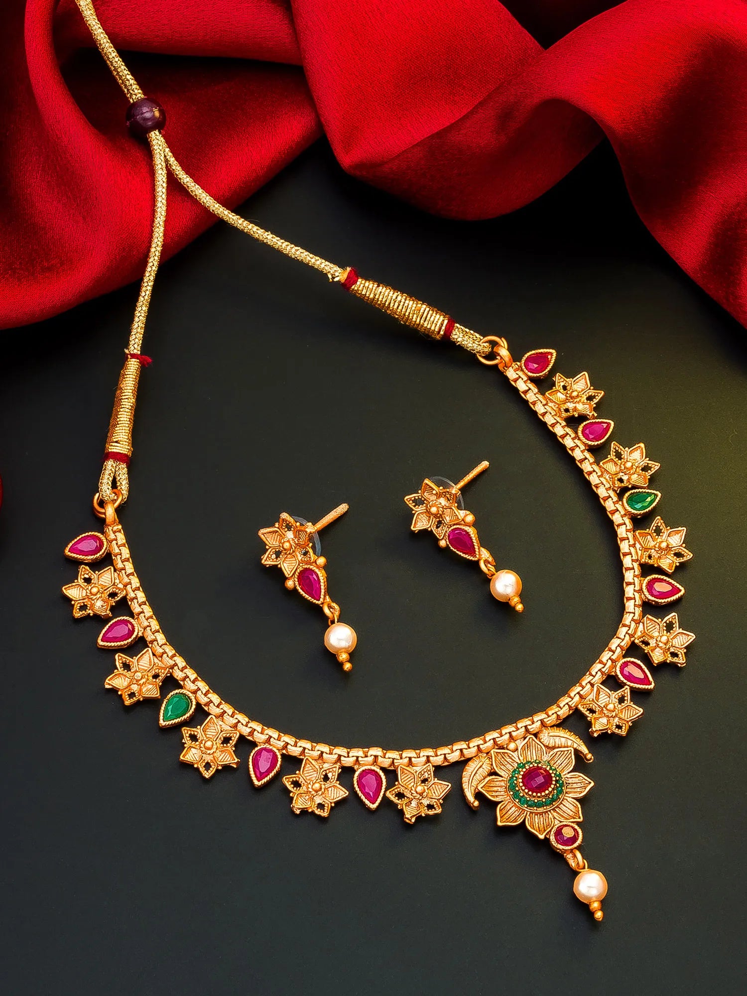 Gold-toned traditional necklace and matching drop earrings with red and green stone accents and pearl details, arranged on a dark surface with red fabric in the background.