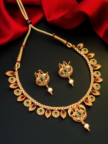 Gold-toned traditional Indian jewelry set featuring a necklace and matching earrings with red and green stone accents and pearl details, displayed on a dark background with red fabric.