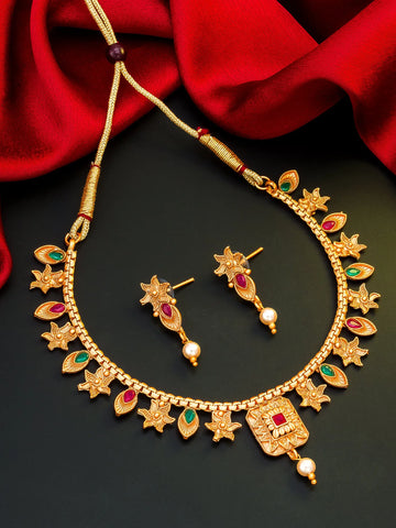 Gold-toned traditional necklace and matching drop earrings with red and green stone accents and pearl details, displayed on a dark surface with red fabric in the background.