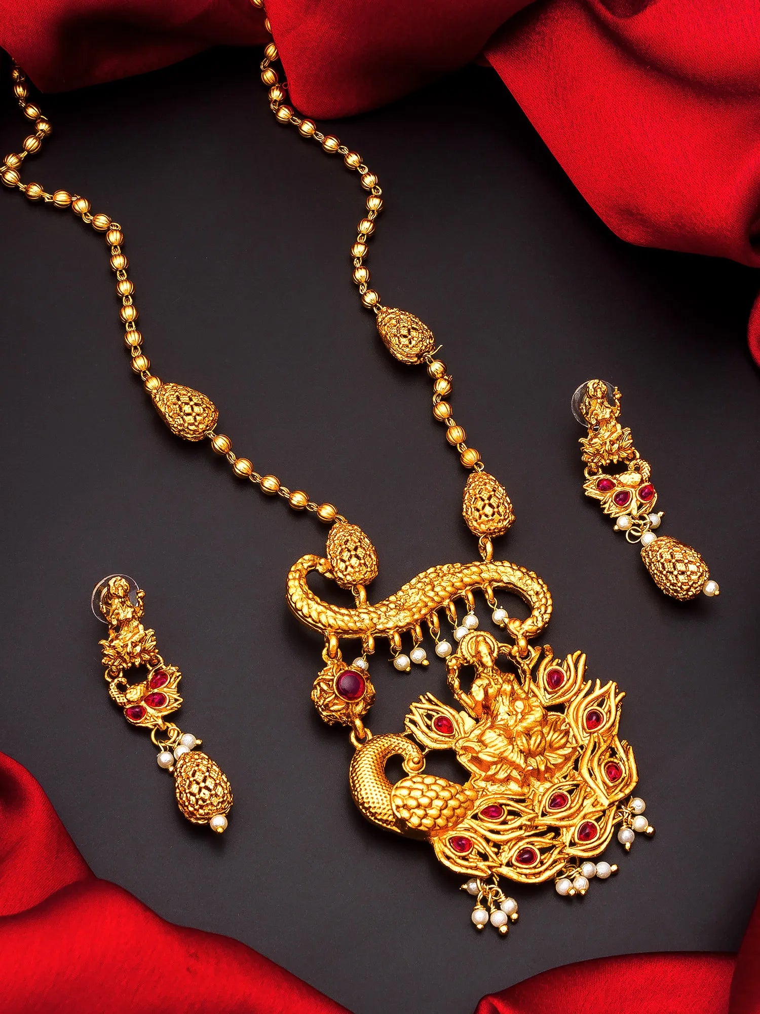 Gold-toned traditional Indian jewelry set featuring a detailed pendant necklace and matching drop earrings, embellished with red stones and small pearl-like beads, arranged on a dark surface with red fabric.