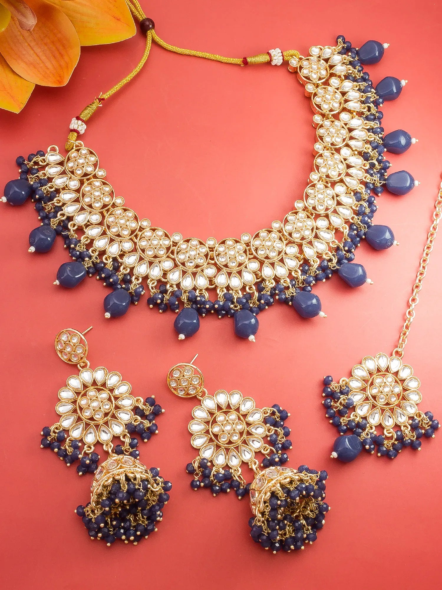 Gold and white stone jewelry set with navy blue bead accents, including a statement necklace, matching earrings, and a chain, arranged on a coral background.