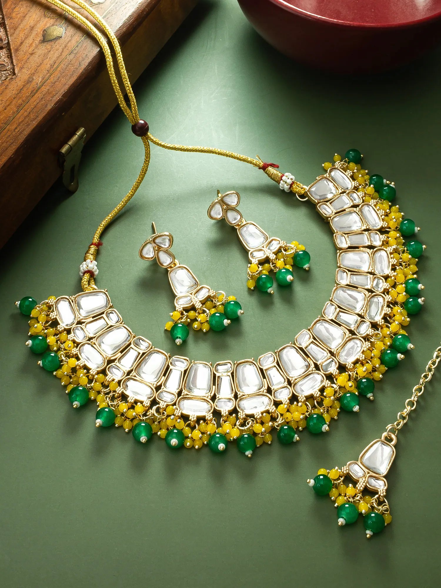 Gold-plated Indian jewelry set featuring a statement necklace, matching earrings, and a maang tikka, embellished with white stones, green beads, and yellow floral accents, displayed on a dark green surface.