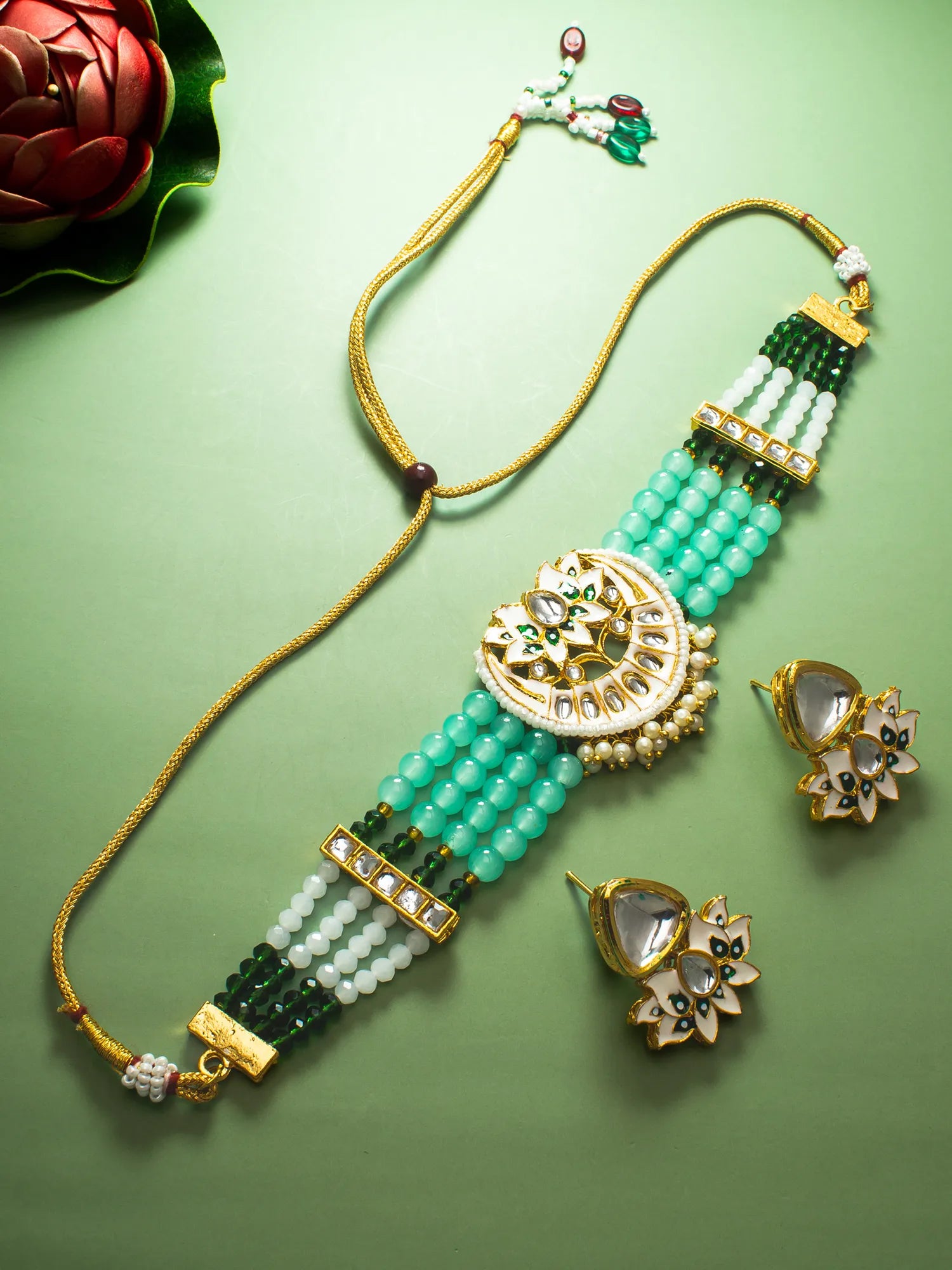 A gold-plated traditional Indian jewelry set featuring a beaded necklace, a matching bracelet, and flower-shaped earrings. The jewelry is decorated with light green and dark green beads, white stones, and gold-plated metalwork, arranged on a solid light green background.