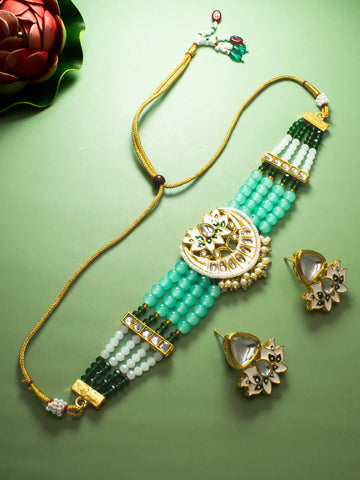 A gold-plated traditional Indian jewelry set featuring a beaded necklace, a matching bracelet, and flower-shaped earrings. The jewelry is decorated with light green and dark green beads, white stones, and gold-plated metalwork, arranged on a solid light green background.
