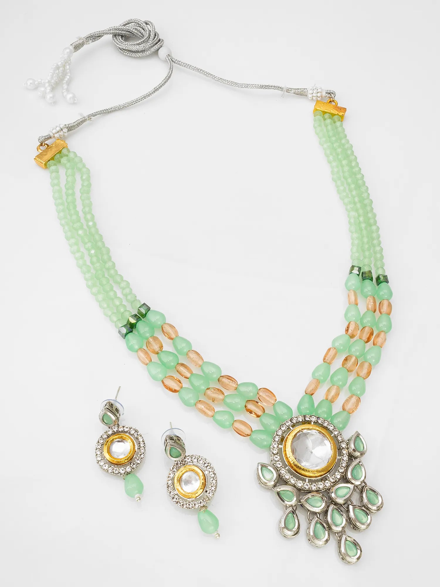 A traditional Indian jewelry set featuring a multi-strand necklace and matching earrings. The necklace has light green beads, peach-colored accent beads, and a central silver-toned pendant with a clear stone and green accents.