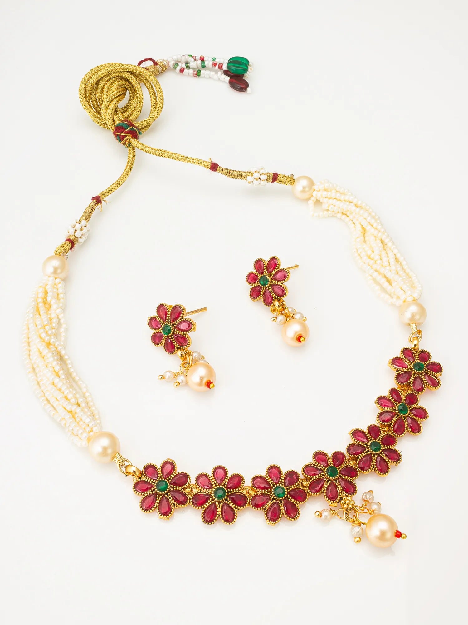 A traditional Indian jewelry set featuring a gold-plated pendant necklace with white pearl strands, red and green beads, and matching gold earrings, displayed on a draped white satin fabric background.