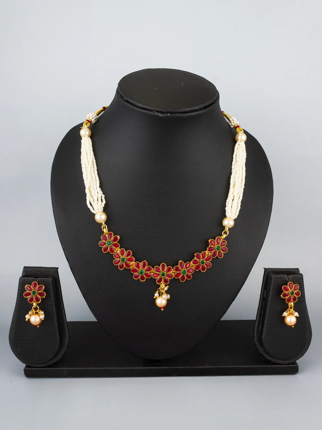 A traditional Indian jewelry set featuring a gold-plated pendant necklace with white pearl strands, red and green beads, and matching gold earrings, displayed on a draped blue satin fabric background.
