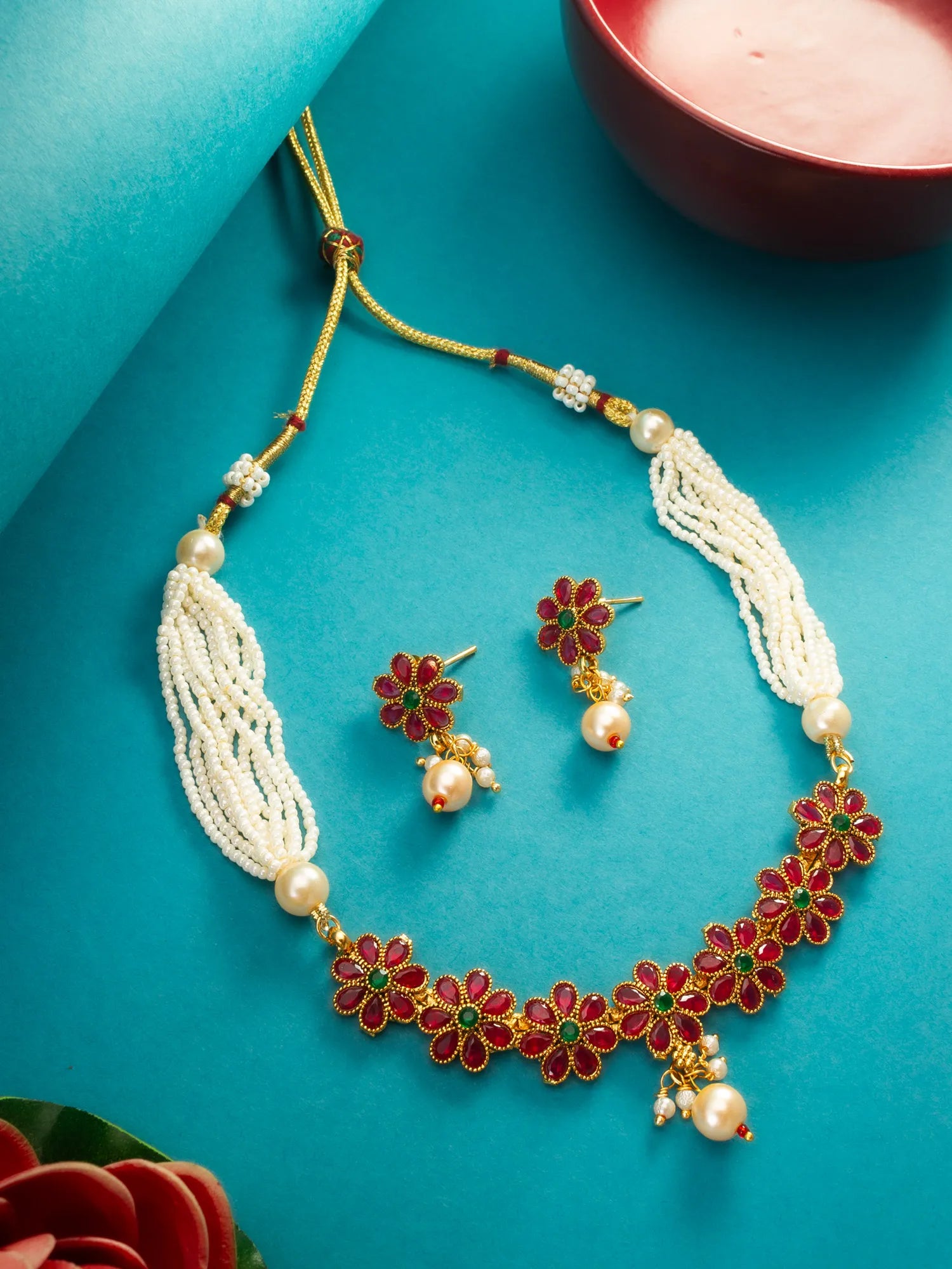 A traditional Indian jewelry set featuring a gold-plated pendant necklace with white pearl strands, red and green beads, and matching gold earrings, displayed on a draped blue satin fabric background.