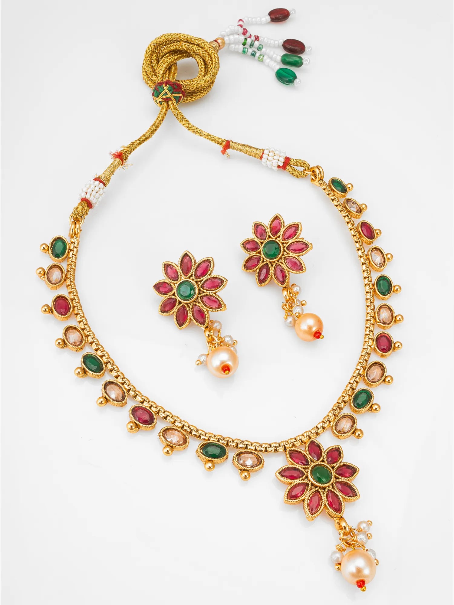 Gold-plated Indian jewelry set featuring a necklace, earrings, and a beaded cord accessory, decorated with red and green stones and white pearls on a white background.