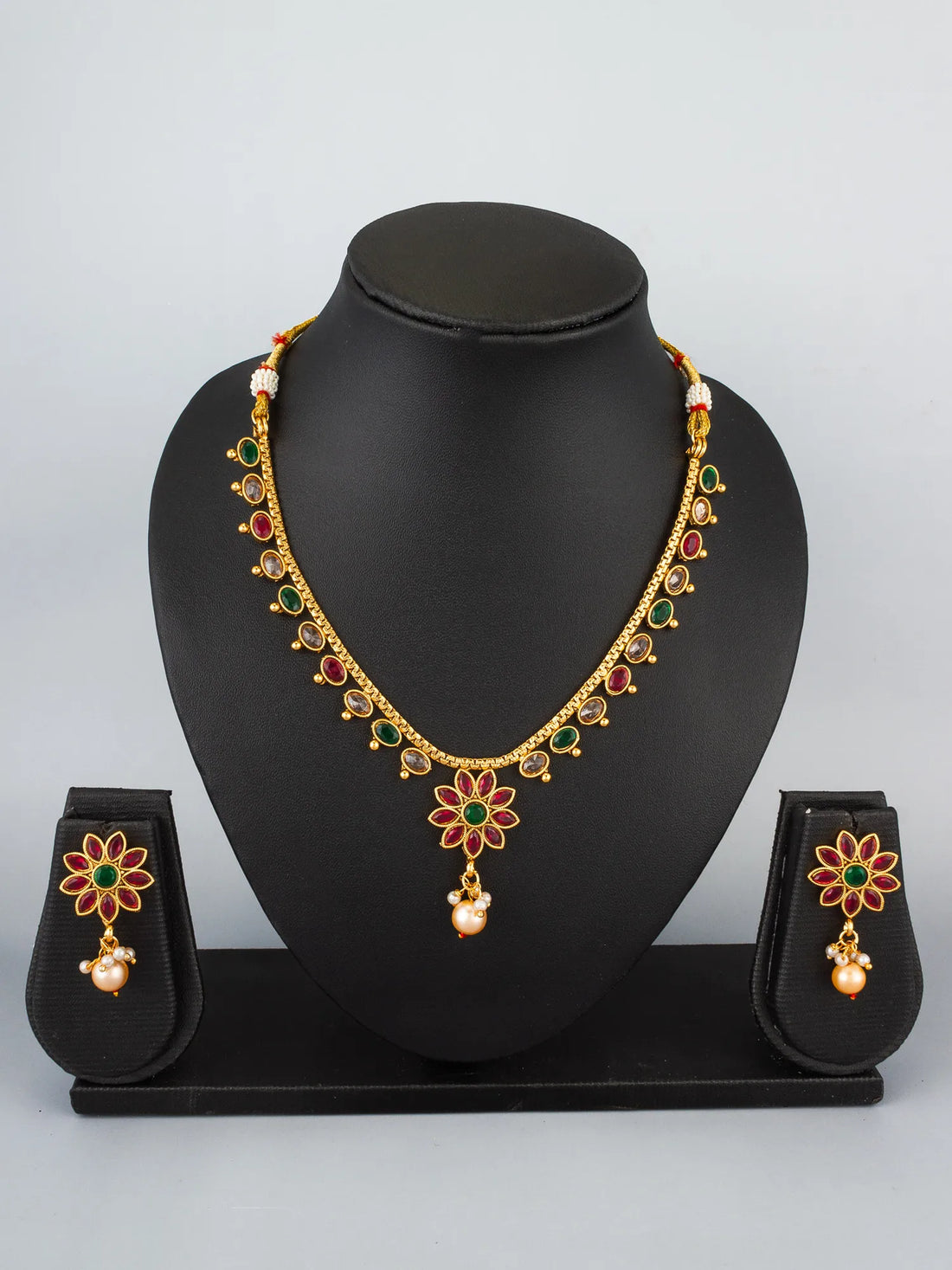 Gold-toned Indian jewelry set featuring a necklace and matching earrings with floral motifs, red and green enamel details, and pearl-like drops, arranged on a bright turquoise background with decorative flowers.