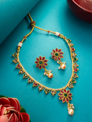 Gold-toned Indian jewelry set featuring a necklace and matching earrings with floral motifs, red and green enamel details, and pearl-like drops, arranged on a bright turquoise background with decorative flowers.