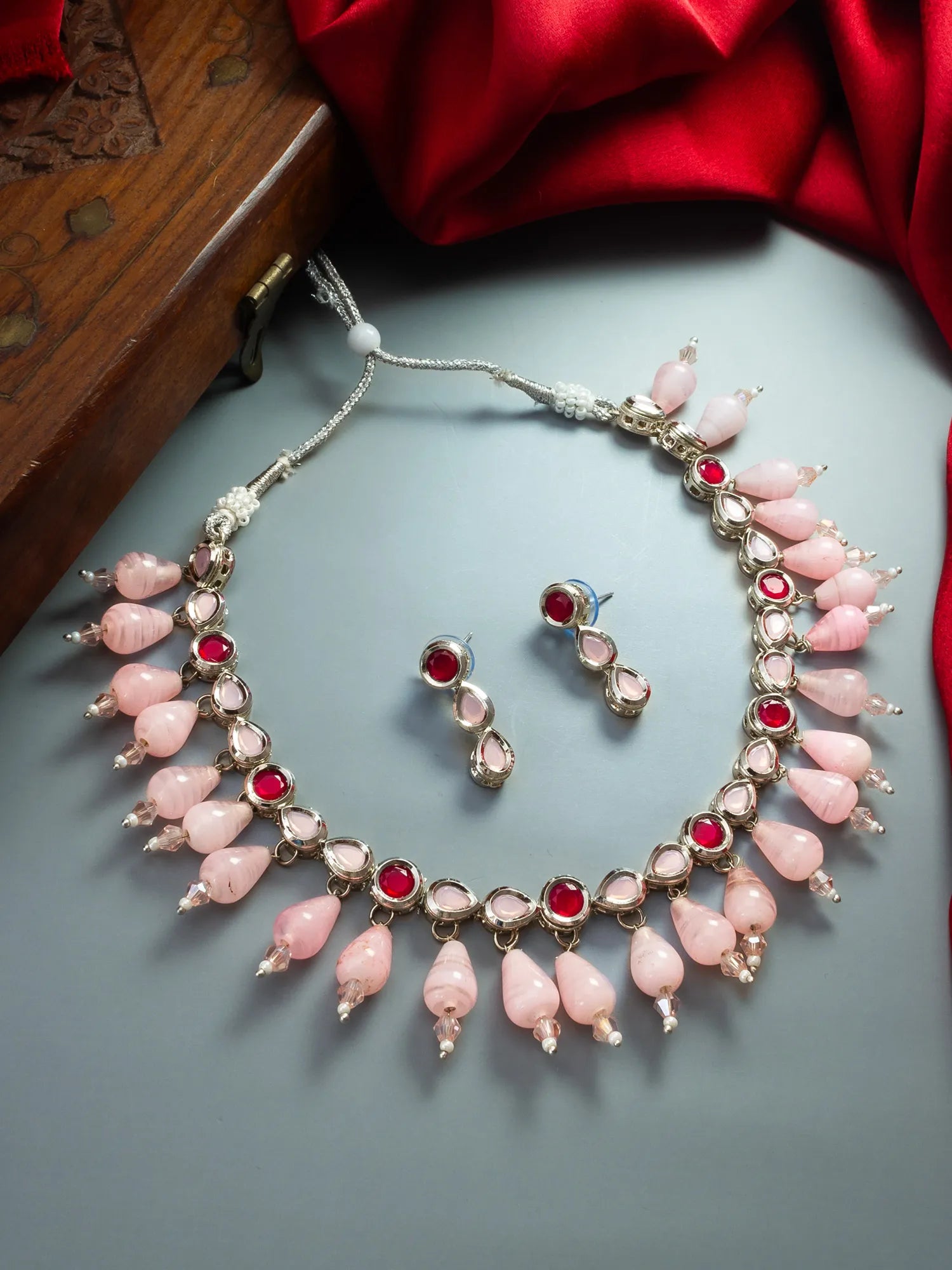A pink and silver jewelry set featuring a statement necklace and matching earrings, decorated with pink teardrop beads, red gemstones, and silver-toned metal, displayed on a gray surface with red fabric and a wooden box in the background.
