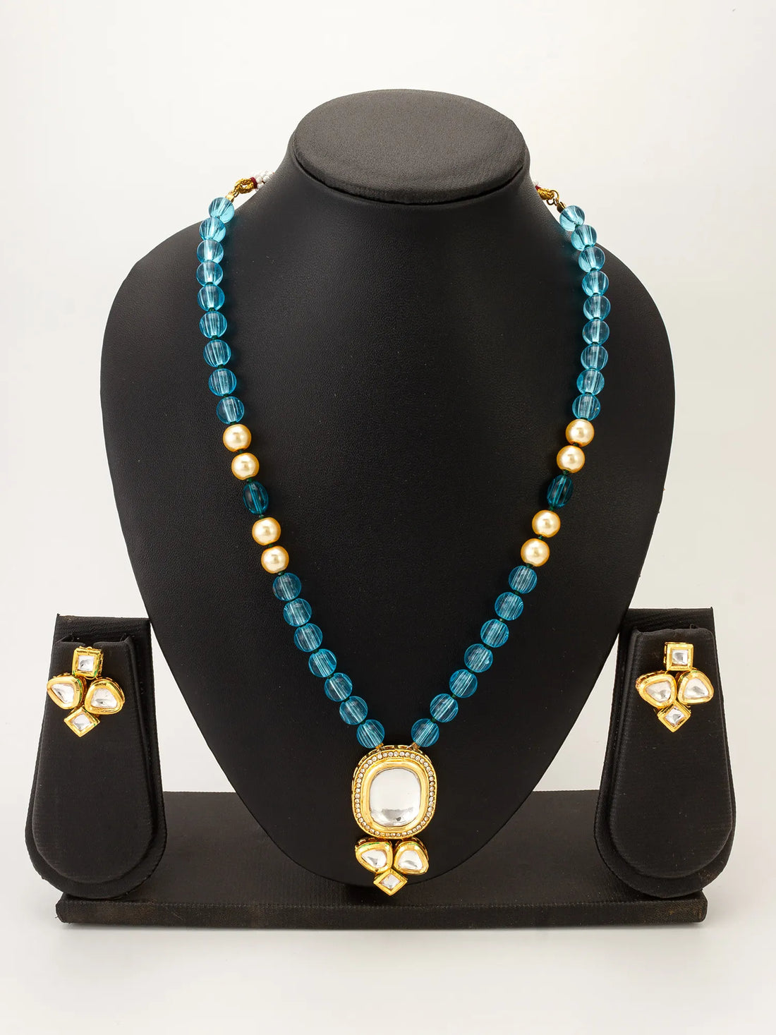 A gold-plated necklace and matching earrings set with faceted blue beads, small gold pearls, and a large oval white stone pendant, displayed on a draped deep burgundy satin fabric.