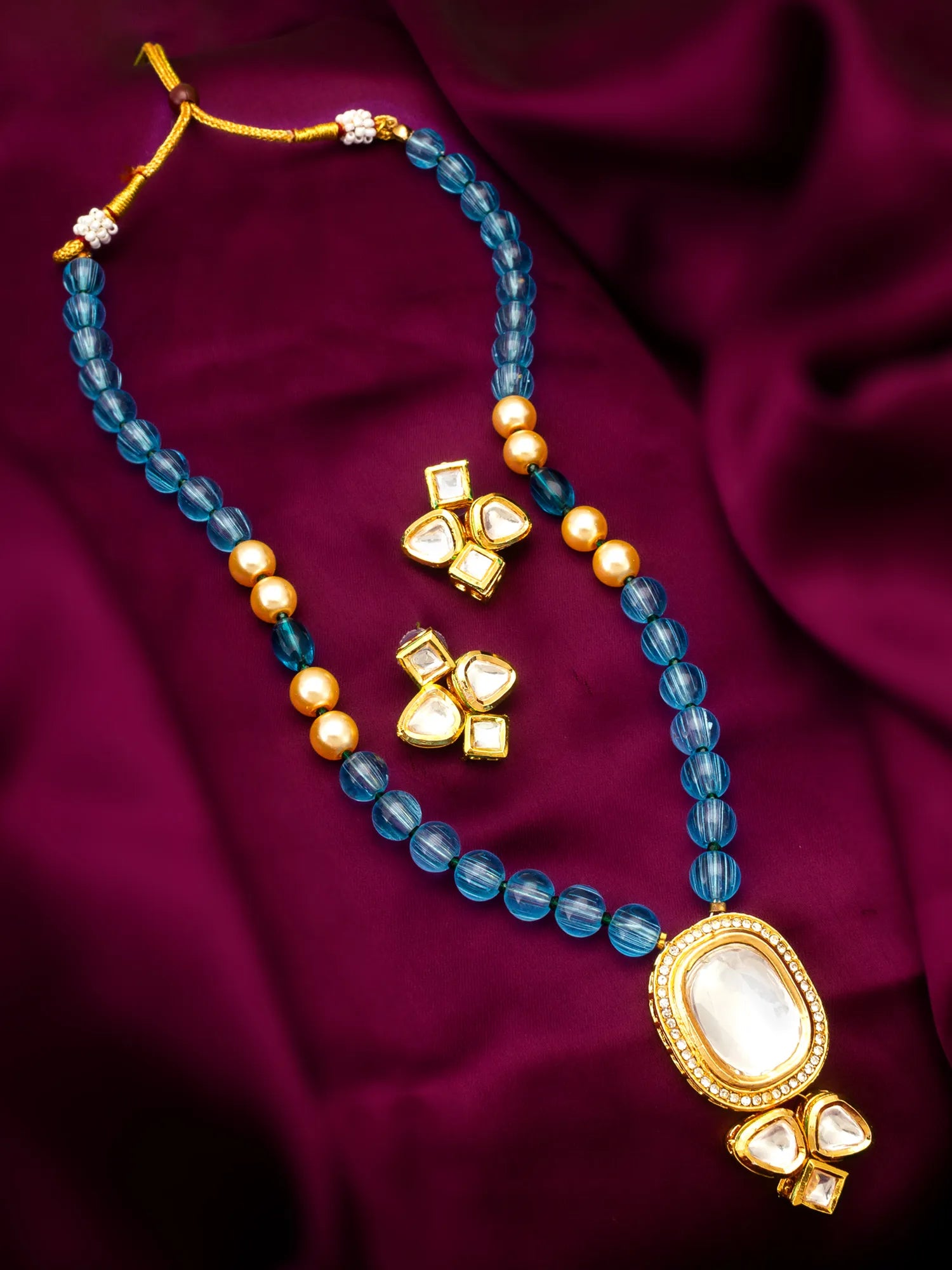 A gold-plated necklace and matching earrings set with faceted blue beads, small gold pearls, and a large oval white stone pendant, displayed on a draped deep burgundy satin fabric.
