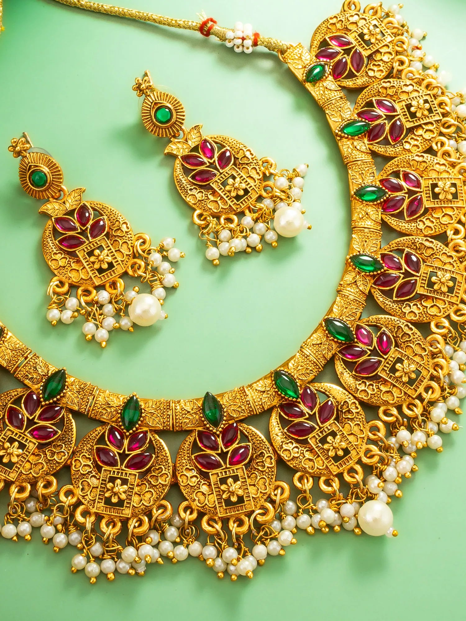 Gold necklace with red and green gemstones and pearl accents on a light green background