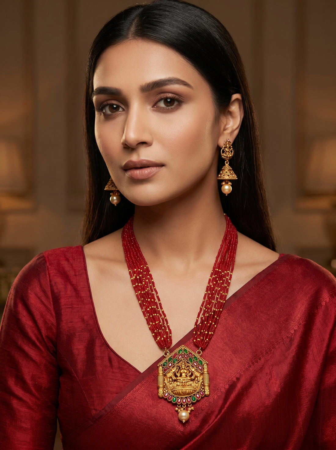 A woman wearing a red traditional garment and a large ornate gold necklace with a detailed pendant, along with matching gold earrings, set against a warm, softly blurred indoor background.