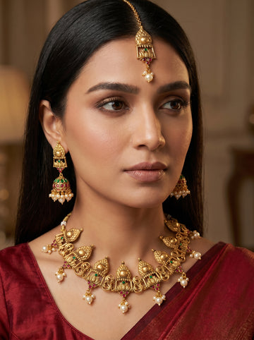 A woman wearing an ornate gold-toned traditional necklace with matching drop earrings and a forehead ornament, styled with a deep red satin garment.