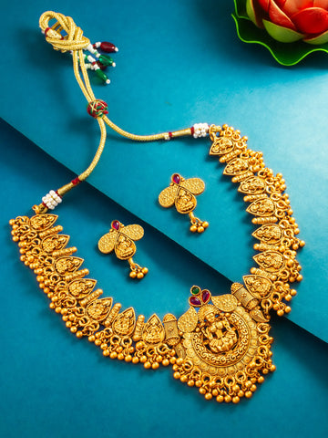 An ornate gold-toned traditional necklace with matching earrings, displayed on a blue background with a small red flower prop in the corner.