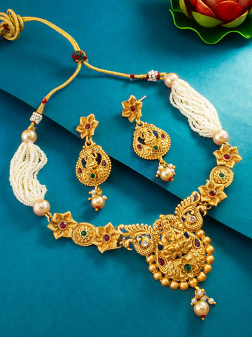 An ornate gold-toned traditional necklace and matching earrings set with pearl-like white strands, floral motifs, and small red and green stone accents, arranged on a teal-blue background.