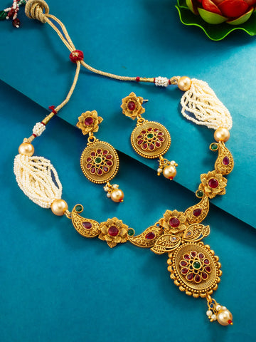 An ornate gold-toned traditional necklace and matching earrings laid out on a teal-blue background. The jewelry features floral motifs, red and green enamel details, white pearl-like beads, and cream tassel ends.