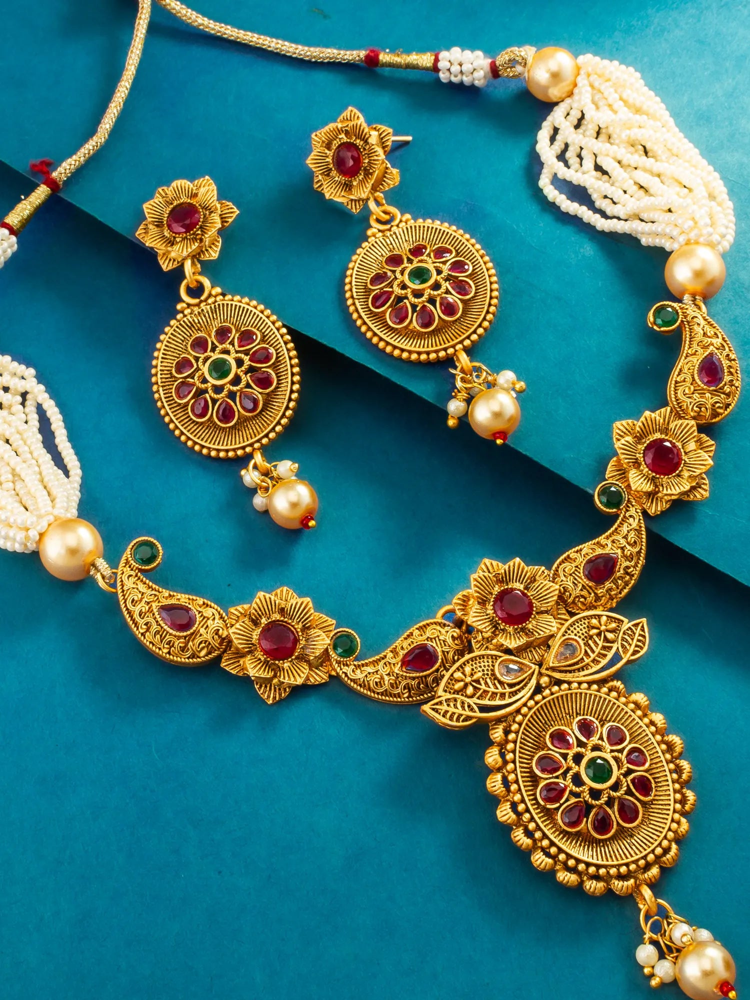 Gold-toned traditional Indian jewelry set including a necklace, earrings, and a bracelet, decorated with red and green stones and white pearl beads, displayed on a teal background.