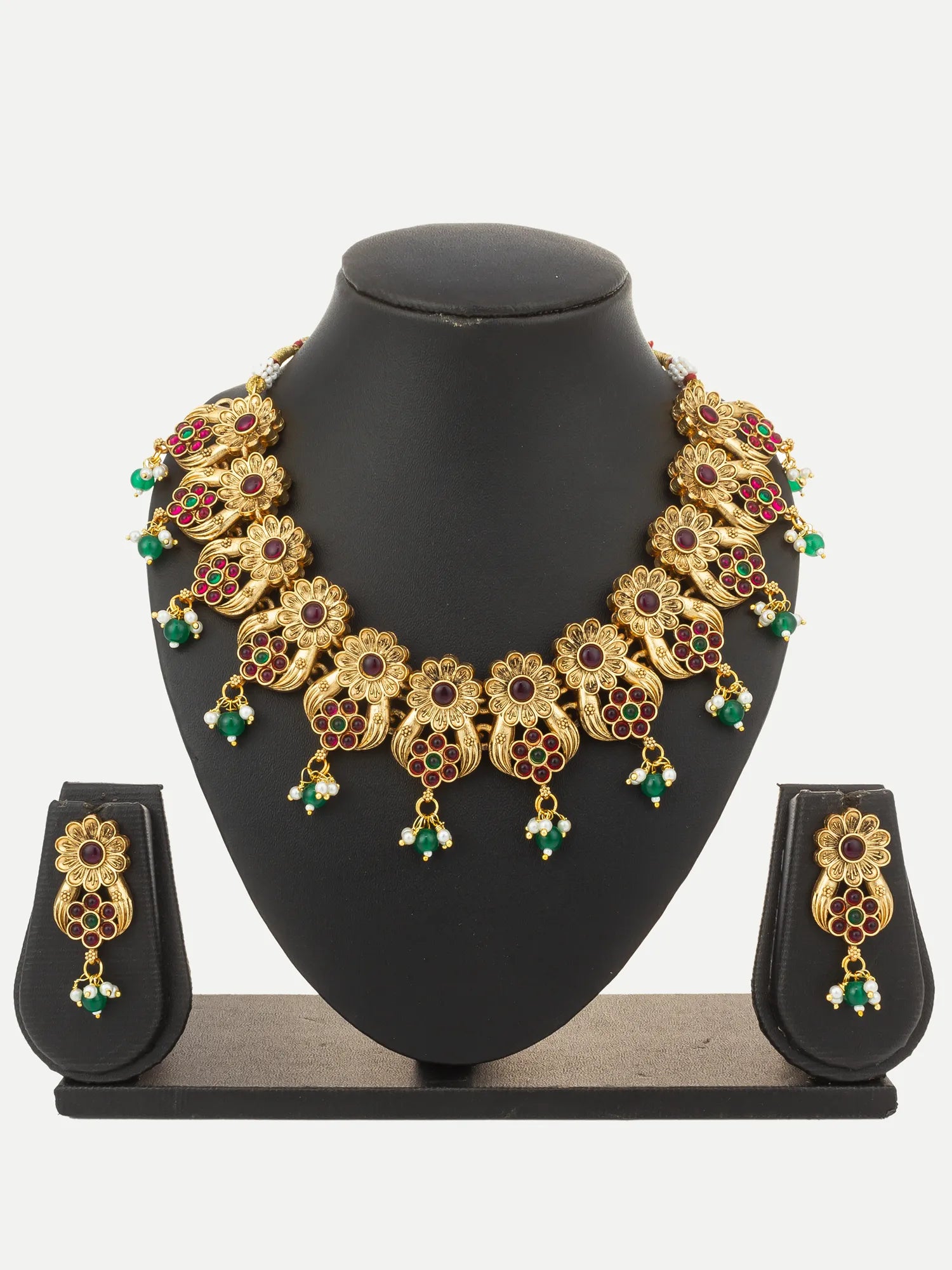 Gold-toned traditional Indian jewelry set featuring a statement necklace and matching earrings, embellished with red and green stones, displayed on a black mannequin bust.
