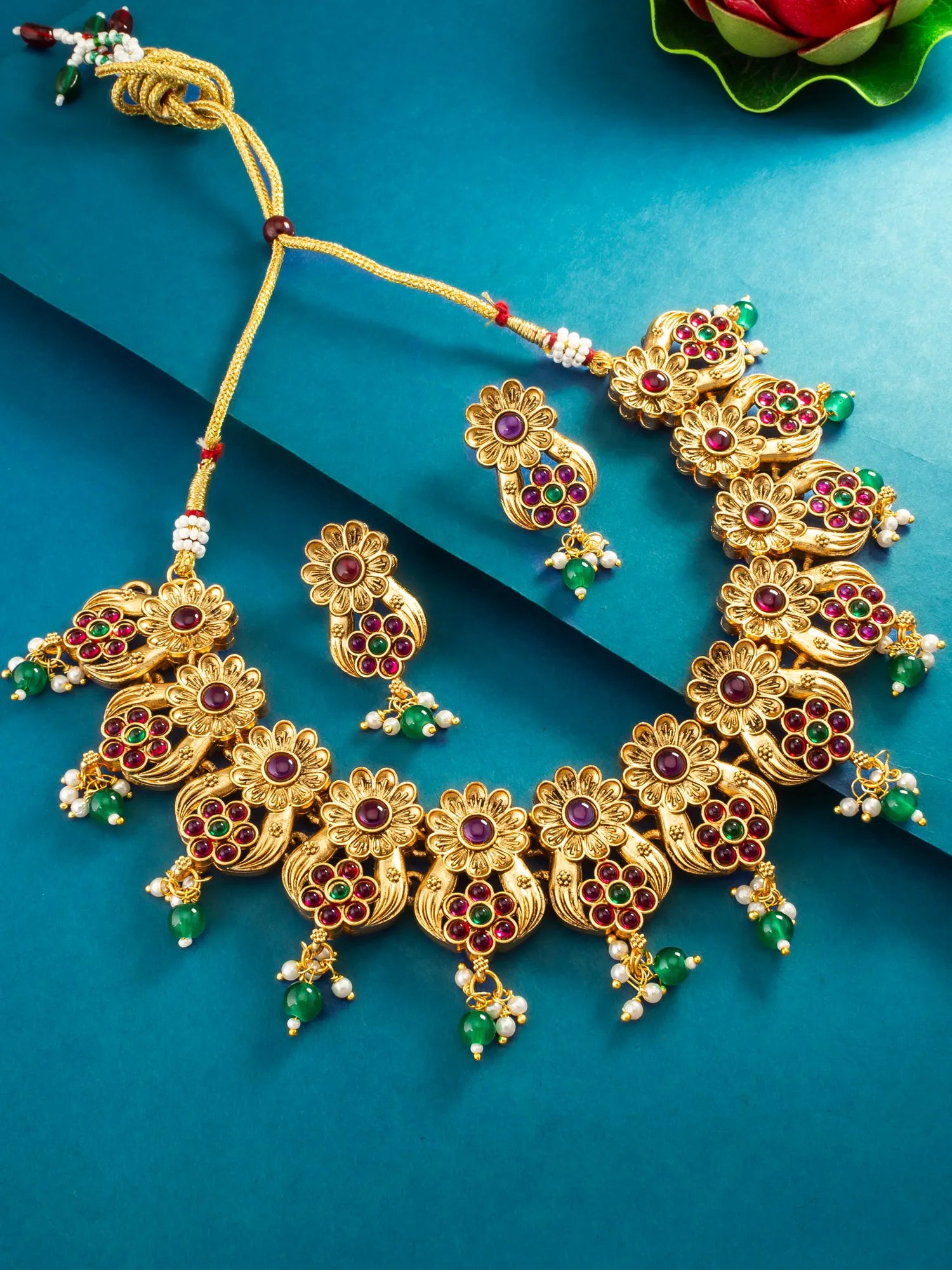 Gold-toned traditional Indian jewelry set featuring a statement necklace and matching earrings, embellished with red and green stones and white pearl-like beads, arranged on a teal-blue background.