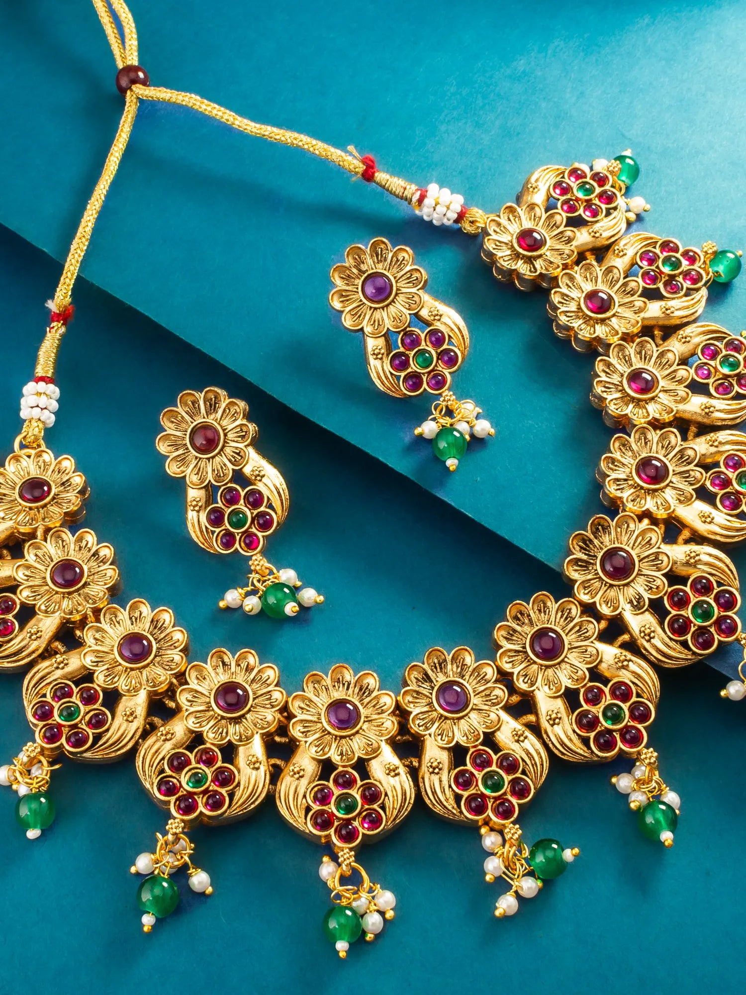 Gold-toned traditional Indian jewelry set featuring a necklace, earrings, and a bracelet, embellished with red and green stones and white pearl-like beads, arranged on a teal background.