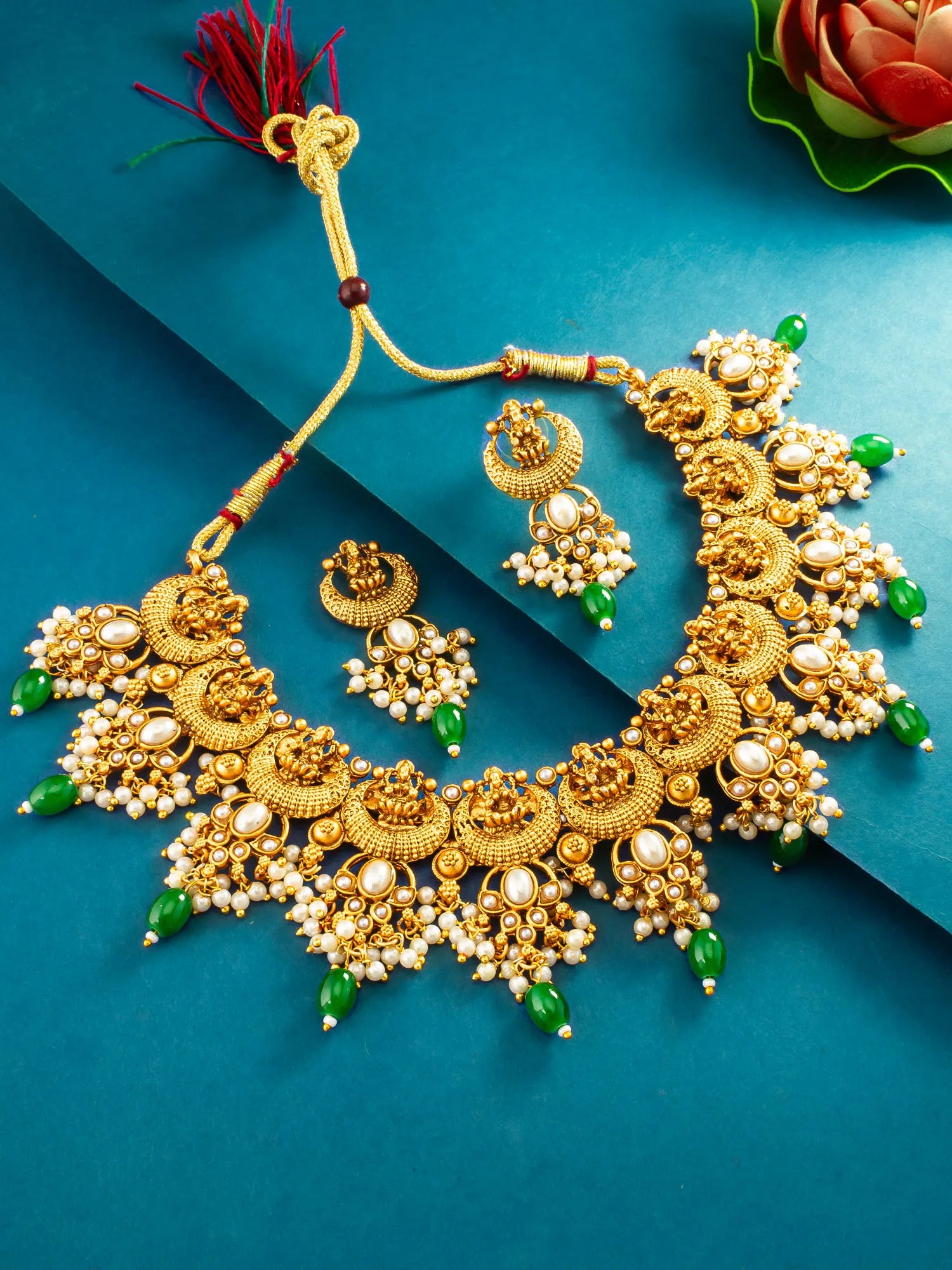 An ornate gold-toned traditional necklace set with matching earrings, decorated with white pearl-like beads, green stone accents, and a red tassel, arranged on a teal-blue background.
